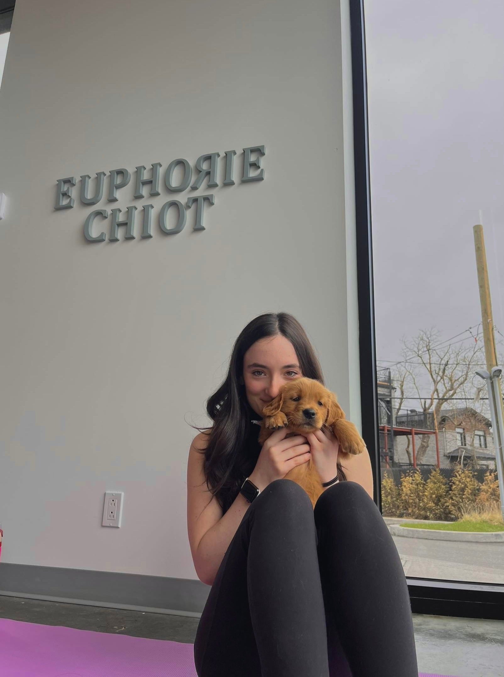 Femme souriante tenant un chiot devant le logo Euphorie Chiot, lors d'une séance de Puppy Yoga sur la Rive-Nord de Montréal. 