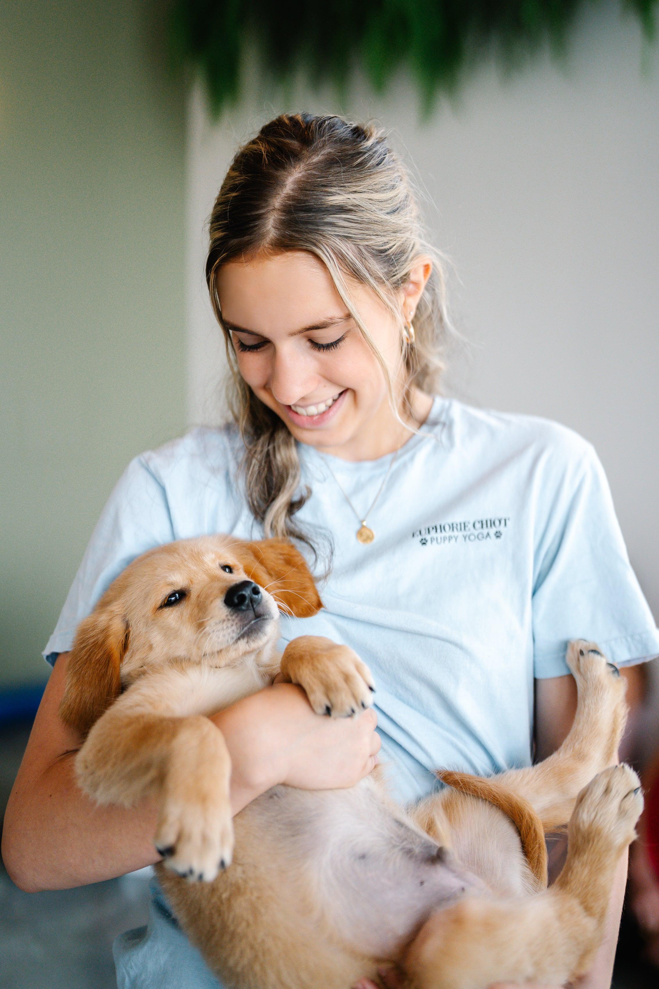 Jeune femme souriante câlinant un chiot Golden Retriever détendu dans ses bras, portant un t-shirt Euphorie Chiot Puppy Yoga.